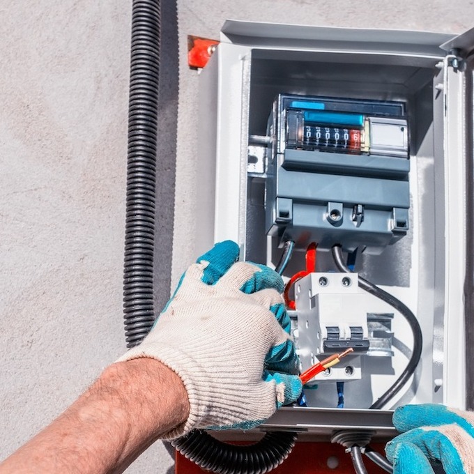 An electrician is tinkering with an electric panel inside an exterior box while wearing gloves.