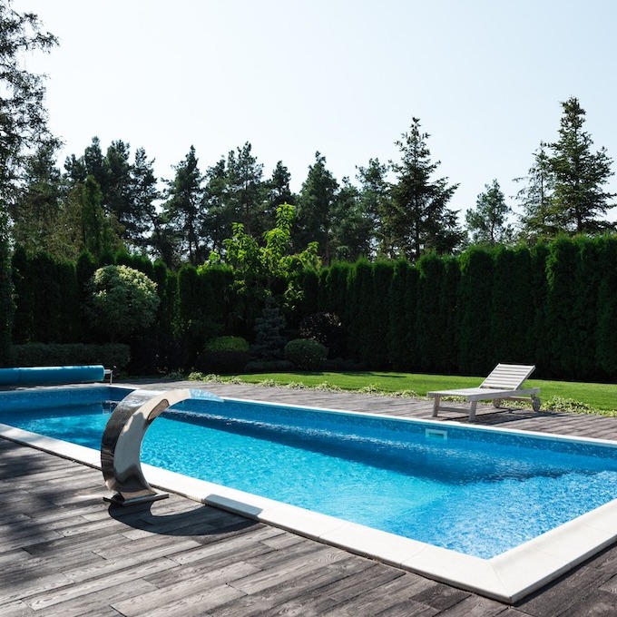 A aquamarine pool with a wooden deck and large trees and shrubs in the background.